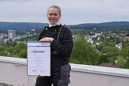 Lehrling des Monats Hanna Keutner hält eine Urkunde in der Hand. Sie steht in Schornsteinfegerkluft auf einem Flachdach im Hintergrund der Ausblick über die Stadt Wiesbaden.