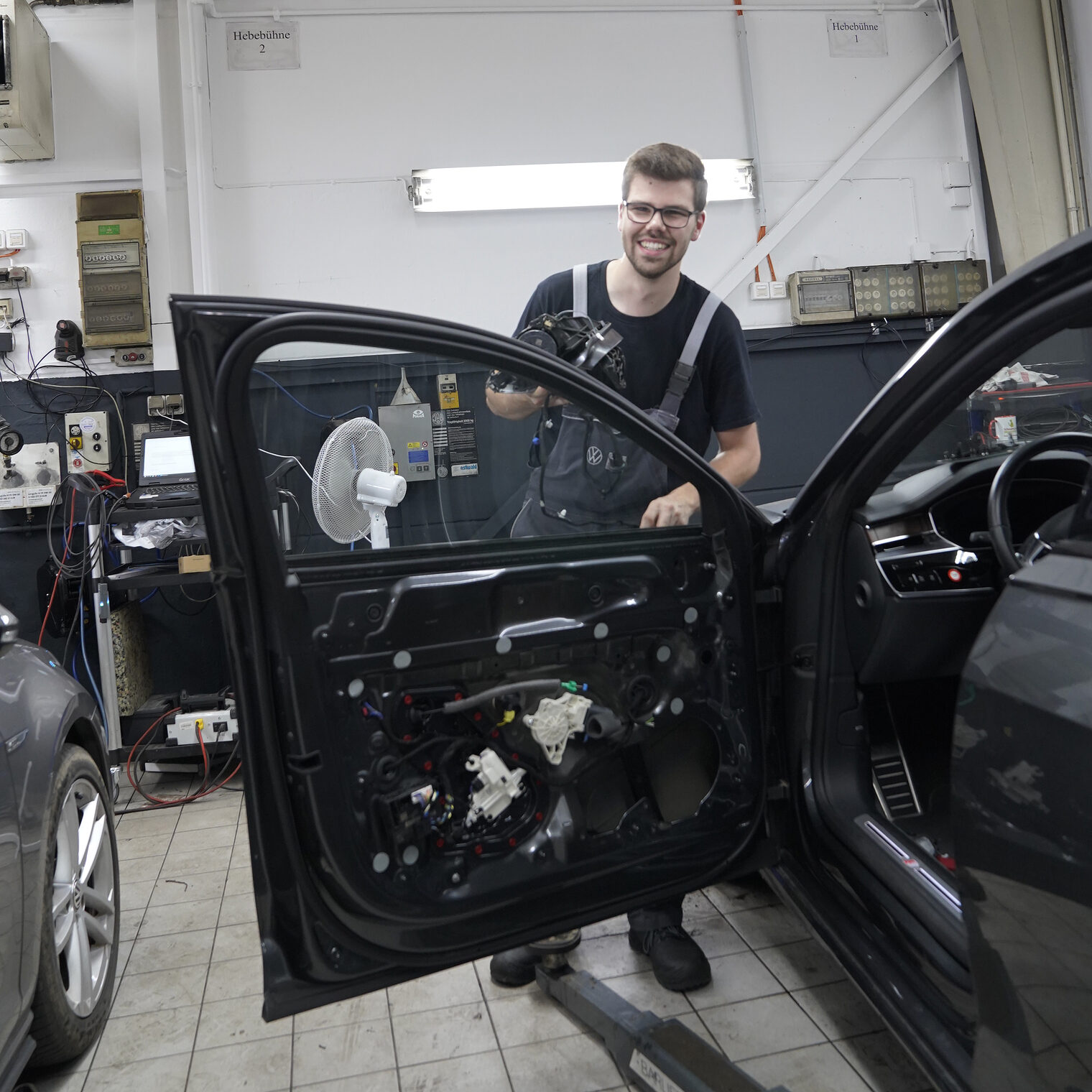 Lehrling des Monats Juli Georg Wahl steht in der Kfz-Werkstatt mit einem Ersatzteil in der Hand hinter einer geöffneten Autotür.